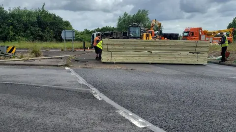 Joshua Brown Overturned lorry on the road- with wooden planks in front of the lorry and two men in High-visibility jacket standing on either side of the wooden planks