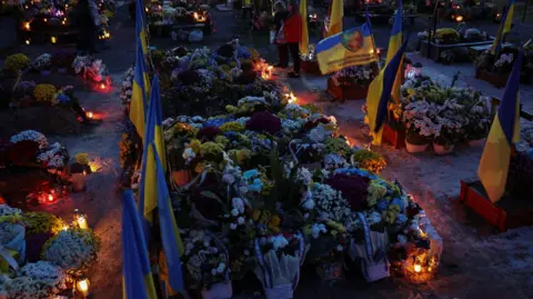 A Ukrainian military graveyard in the dusk. Each grave is covered in flowers and has a Ukrainian flag at the end of it. Candles at the end of each grave provide some light