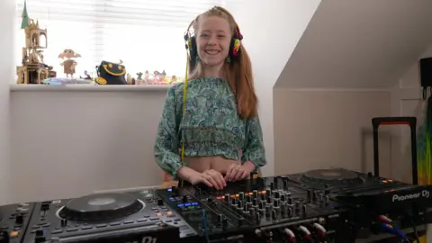 Laura Foster/BBC A young girl with long blonde hair is in her house smiling and stood behind some large black DJ decks full of dials and lights