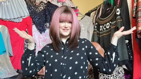 Caroline Butler Caroline in her spare room, full of charity shop items. She is holing her arms out, wearing a pink and brown wig, with a black top with stars on, with clothing all around her. 