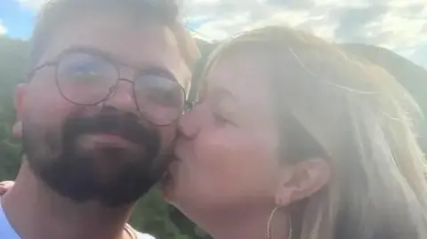 Selfie-style image of a man with a beard and wearing glasses being kissed by a woman with blonde hair.