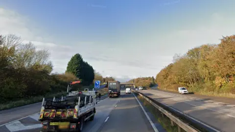 Google Vehicles travelling on a motorway that has two lanes. A slip road can also be seen on the far left side. There are trees lining the side of the carriageways and a concrete barrier separates the two sides of the road. The sky in the background is blue.  