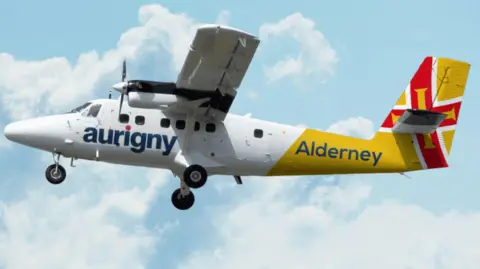 The image shows a small passenger aircraft in mid‑flight against a bright blue sky with scattered clouds. The plane has a white fuselage with the word “aurigny” written in large blue letters along the side. The rear section is yellow and carries the word “Alderney” in blue text. The tail fin is painted in a bold red, yellow and white pattern.