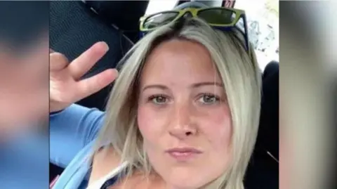 A woman with short blonde hair, wearing a light blue top and sun glasses on her head, and holding one hand up in a peace sign as she pouts and looks at the camera.