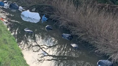 Gavin Mills Bags of rubbish dumped in a river flowing between fields.