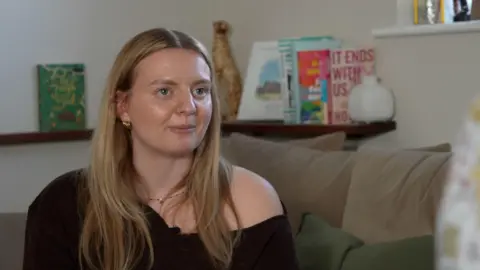 Qays Najm/BBC A woman with blonde hair and blue eyes looks to the side of the camera. She is wearing a black top. Several books are on a shelf behind her.