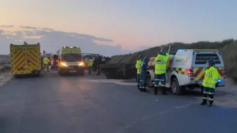 HM Coastguard Coastguard and emergency service workers in high-visibility uniforms are seen near their vehicles on a tarmac road by the grassy beach dunes at sunset.