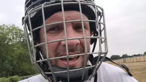 Will Chadwick as a close up of his fac wearing a skate helmet and with an open mouthed smile at the camera. Behind him are grass and trees.