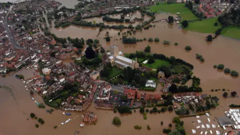 An aerial view showing Tewkesbury during widespread flooding. Brown floodwater are seen on most of the town, with the church one of the few places not flooded