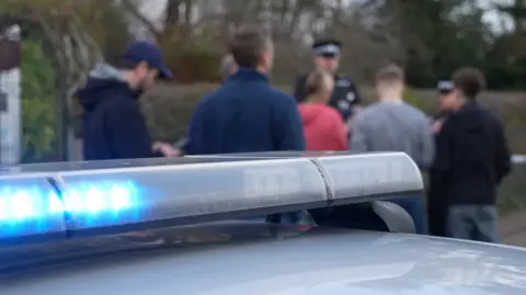 Qays Najm/BBC A blurred image of a group of people, their backs to the camera, facing a police officer. In the foreground is the blue light of a police car