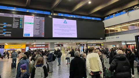 Passengers stand and wait in the main concourse at London Euston station beneath departure boards showing delays and cancellations, with shops and platforms visible in the background.