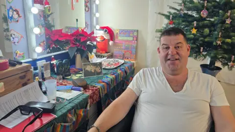 Hayley Coyle/BBC Damian Williams sat in his dressing room wearing a white tshirt with a Christmas tree in the background and his glasses, bubble bath, framed photos and a lighted mirror beside him