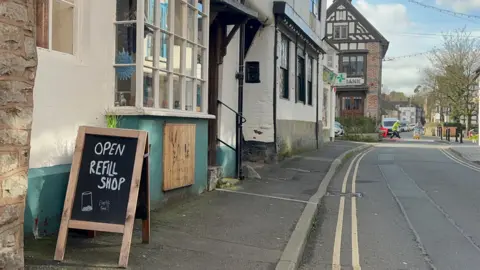 ELLEN KNIGHT/BBC The exterior of Sam's shop can be seen, from a side-on angle, looking down the pavement. In the foreground is a black chalkboard with 'Open Refill Shop' written on it in white letters. The outside of the shop is whitewashed, with a teal blue border at the bottom. There is a large bay window, and underneath it is a wooden sign that reads 'The Simple Life Unwrapped'.