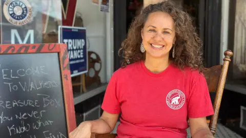 Adele Walker sits in a chair nearby a Trump-Vance campaign sign 