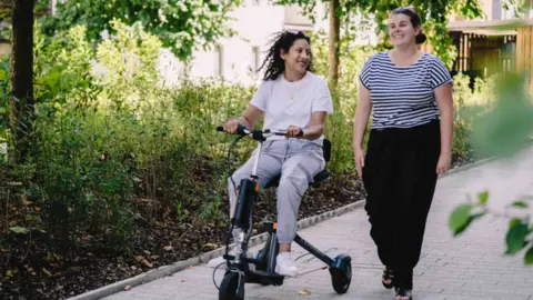 Supersmith A woman riding a three wheeled scooter and talking with another woman who is walking by her side