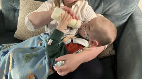 Deborah Rowe sitting on a grey sofa, with her son Jonah lying on her lap with a blue blanket. She is feeding him a bottle of milk. He has a nasogastric tube inserted and is lying against his mother's crooked arm.