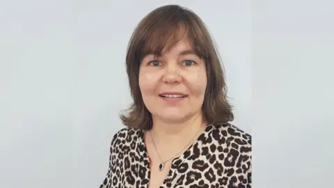 University of Salford Daiga Kamerāde looking straight down the camera against a white background. She has brown, shoulder-length hair and blue eyes. She is smiling and her teeth are showing. She is wearing an animal-print blouse and a pendant necklace.