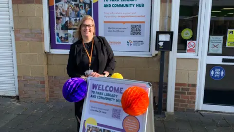 Helena Robinson stands outside the new hub, just behind a new sign board which says "Welcome to Park Library and Community Hub." Different coloured pom poms are stuck to the board.