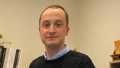 Jordan Wallace A young man with fair hair looking at the camera. He is wearing a light blue shirt under a dark round-neck jumper