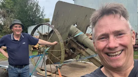 Ian Rogers and Taff Gillingham with a rusty World War One Howitzer gun on a trailer. The gun is in the middle of them and has been strapped to the trailer. 