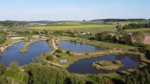 An aerial photograph taken of green fields with hedges and trees under a blue sky. Three small lakes are in the centre of the picture with stretches of land linking them. A roadway with a car on it runs around the centre lake. 