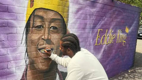 BBC A man kneeling down beside the mural on a long breezeblock wall. The artist is adding a gold tooth with a metallic gold pen. The mural shows Eddie Kinuthia smiling, wearing a yellow cap and black jacket. Beside him is scrawled writing, which reads 'Eddie King' on the right and 'talent, godsent, I'm chosen' on the left, with various music notes against a bright purple background.