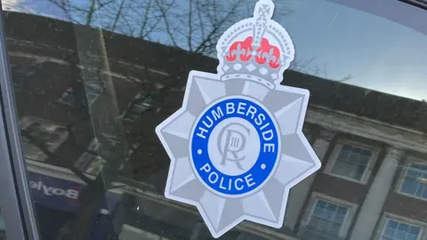 BBC A close-up of a Humberside police badge on the window of one of their vehicles. It is a grey star, with a blue circle in the centre and is topped with a grey and red crown.
