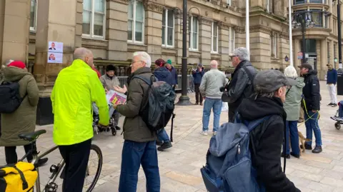 Alexander Brock Men and women stand outside the council house entrance, some with leaflets in their hands. One man in a fluorescent jacket holding a bike's handles is standing talking to another man. There is another man with a dog. 