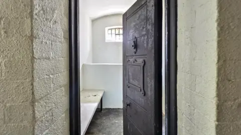 Paul Fish Auctions An imposing heavy grey metal cell door stands ajar, a bed can just be seen inside.