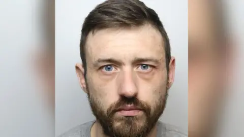 A man with brown hair and beard, with grey eyes staring at camera.