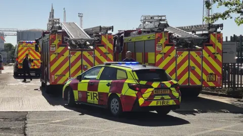 Four red and yellow fire engines with ladders on the roof, and a fire response car with flashing lights on the roof, are parked. There is a metal fence to the right.