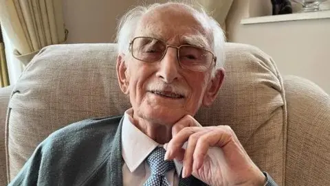 Man wearing glasses looks straight into the camera lens. He's smiling and wearing a white shirt with a blue and white spotted tie and grey cardigan