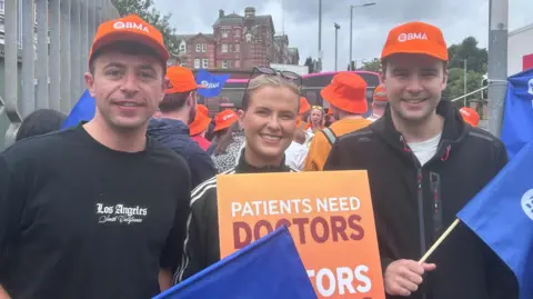 Orla Murray standing with two other protesters holding blue BMA flags and wearing orange hats dressed in black. Orla has blonde hair, is wearing glasses on her head as well as an adidas top