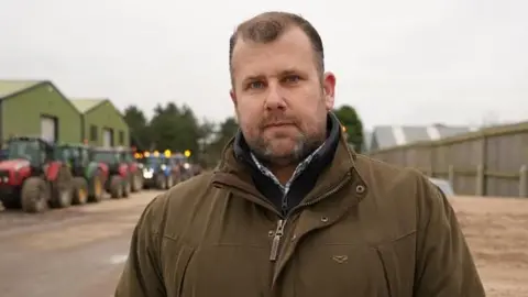 Shaun Whitmore/BBC A man with short brown hair smiles at the camera. He is wearing a green coloured coat with a navy jumper underneath. Behind him tractors can be seen lined up behind one another.