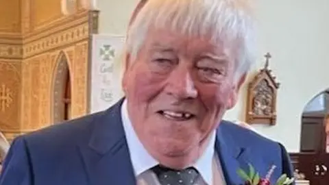 A man with grey hair and wearing a blue suit, white shirt and grey tie stands inside a church. 