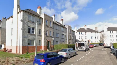 A street of three storey flat buildings under a blue cloudy sky with vehicles parked on the road in front