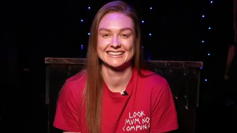 Jodie Bartle is sitting in a dark room which has blue fairy lights hanging on the wall and a table. Bartle has long hair and she is wearing a red T-shirt.