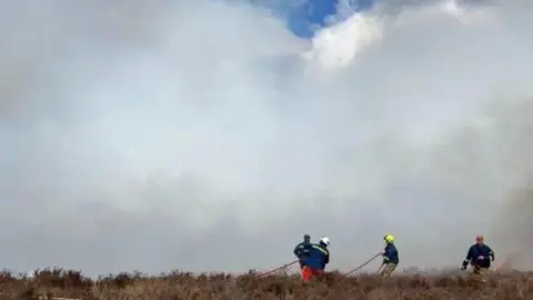 staffordshire Fire The backs of four figures wearing blue protective jackets and helmets can be seen. They are standing in brown, parched looking grass as smoke can be seen the other side of them.