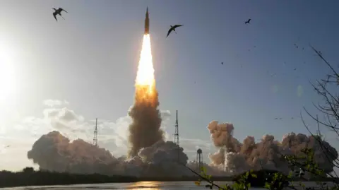 Artemis rocket launches off with bright flames, smoke and the silhouettes of birds in the foreground