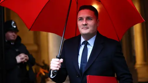 Wes Streeting in the rain with an red umbrella. He is wearing a suit and walking 