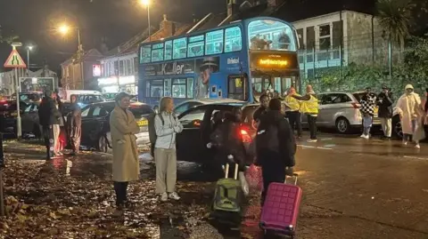 Residents wearing winter coats and carrying suitcases stand outside in a busy street looking towards the building. It is dark, and there is a bus stopped in the middle of the street.