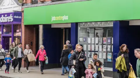 People, including families, walking on a street past the Job Centre Plus employment office in Chester. The street is busy, the weather is dry but people are wearing coats. 