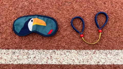 One eye mask and a teder were placed on a running track. There is a picture of a token on it in the mask. The ther has a small yellow strap with a blue loop at each end. 