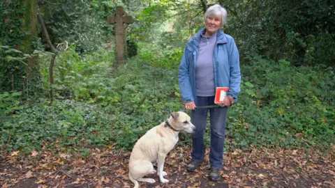 Volunteer Judith Thompson, wearing a blue jacket and blue jeans, stands with a light coloured dog in an overgrown graveyard
