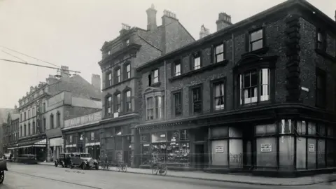 Wirral Libraries Black and white image of Argyle Street looking towards the Scala Cinema. There are old motor cars and bicycles parked on the side of the street
