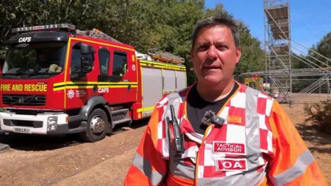 Matt Rowe has short dark hair and is wearing an orange hi-vis jacket. Behind him is a fire engine.