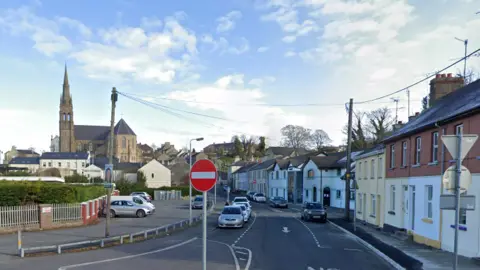 A screengrab of a Google Maps image of Stream Street in Downpatrick. Terraced houses of different designs line one side of the street and a number of cars are parked in parking bays. There is a large church on a hill on the left side of the image and a red "no entry" road sign in the middle of the picture. 