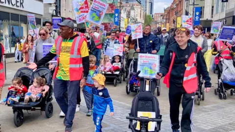 Nicola Rees/BBC A crowd if people walk down a high street towards the camera. People are wearing colourful clothes and neon high-vis jackets, many of them pushing prams or holding hands with children, and others holding placards with slogans about paternity leave.