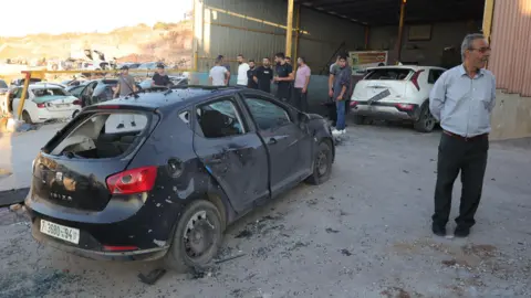 EPA Palestinians stand near a car damaged in an Israeli air strike in Tubas, in the occupied West Bank (5 September 2024)