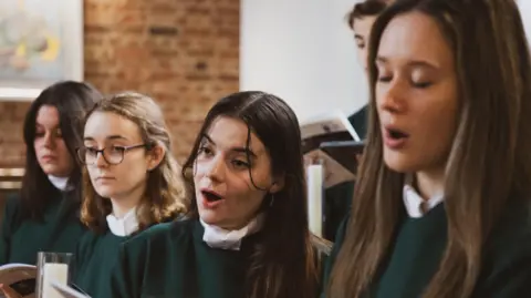 Singers from the University of Exeter's chapel choir performing a song. Four young women are in the front row wearing green tunics. A couple of male singers wearing the same clothes are behind them.